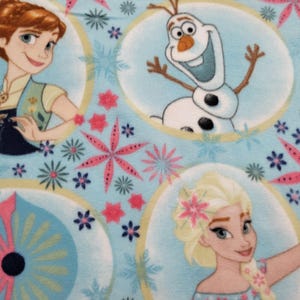May include: Light blue fabric featuring cartoon characters Anna, Elsa, and Olaf from the movie Frozen. The characters are in oval frames, surrounded by pink and blue floral and snowflake patterns. The fabric has a soft, textured appearance.
