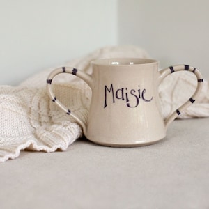 May include: A beige ceramic cup with two handles, decorated with black stripes. The cup has the name "Maisie" written on the front in black.