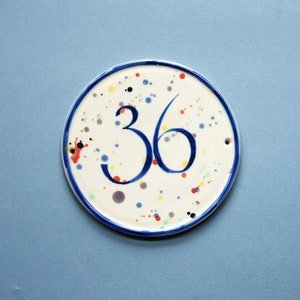 May include: A white ceramic round sign with a blue border and colourful polka dots. The number 36 is written in blue cursive.
