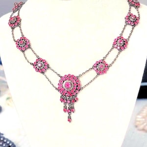 Pode incluir: Um colar vintage com corrente prateada e detalhes em pedras preciosas rosa. O colar apresenta um design floral com um pingente central e gotas em cascata. As pedras são cravejadas em um padrão delicado e ornamentado.