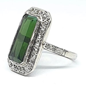May include: A silver ring featuring a large, rectangular, faceted, emerald-green gemstone. The gemstone is surrounded by a border of small, clear gemstones. The ring's band is also detailed with small gemstones.