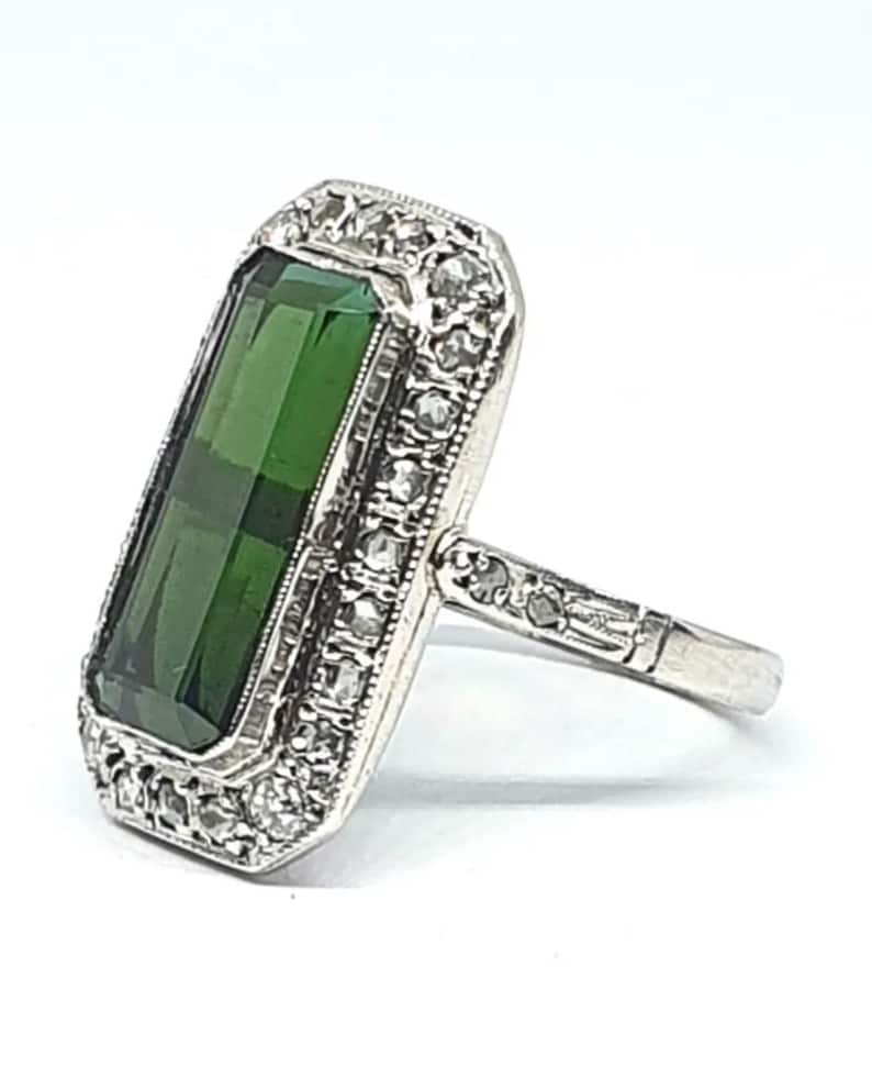 May include: A silver ring featuring a large, rectangular, faceted, emerald-green gemstone. The gemstone is surrounded by a border of small, clear gemstones. The ring's band is also detailed with small gemstones.