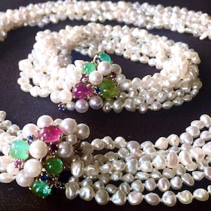 Può includere: Un set di collana e bracciale di perle a più fili. Le perle bianche sono impreziosite da gruppi di pietre preziose colorate, tra cui pietre rosa, verdi e blu. I gioielli sono esposti su una superficie scura.
