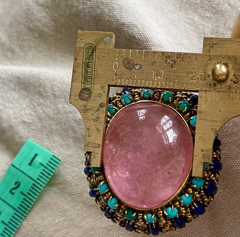 May include: A pink gemstone ring with a gold setting and a border of blue and turquoise beads. The ring is being measured with a ruler, showing the stone is approximately 1.5 inches in diameter.