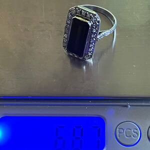 May include: A silver ring with a rectangular black stone, surrounded by small clear stones. The ring is on a scale, with a digital display showing a weight of 6.87.