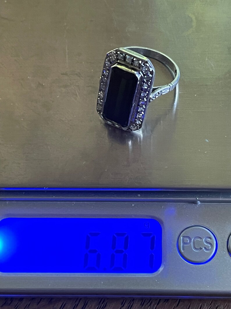 May include: A silver ring with a rectangular black stone, surrounded by small clear stones. The ring is on a scale, with a digital display showing a weight of 6.87.