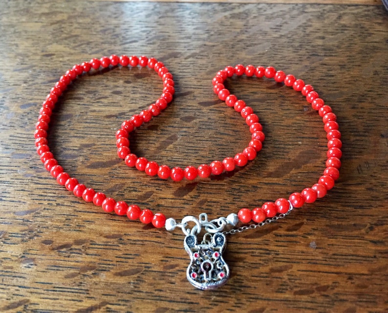 Puede incluir: Un collar de cuentas rojas con un colgante de candado plateado. El candado tiene detalles intrincados y joyas rojas.