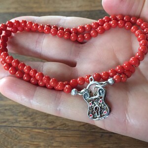 Puede incluir: Un collar de cuentas de coral rojo con un colgante de candado de plata. El collar es de doble cadena y tiene un aspecto delicado y femenino.