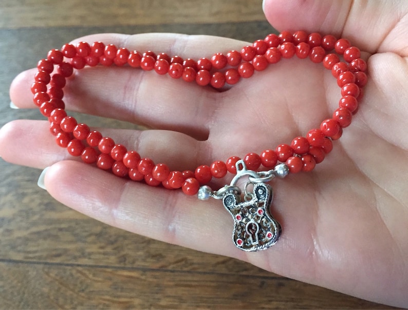 Puede incluir: Un collar de cuentas de coral rojo con un colgante de candado de plata. El collar es de doble cadena y tiene un aspecto delicado y femenino.