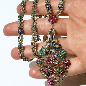 Könnte beinhalten: Silberfarbenes Halsketten- und Armband-Set mit floralen Designs. Die Halskette hat einen Anhänger mit einem Strauß farbiger Edelsteine, darunter rosa, blau, gelb und grün. Armband und Kette haben kleine, farbige Blumenakzente.