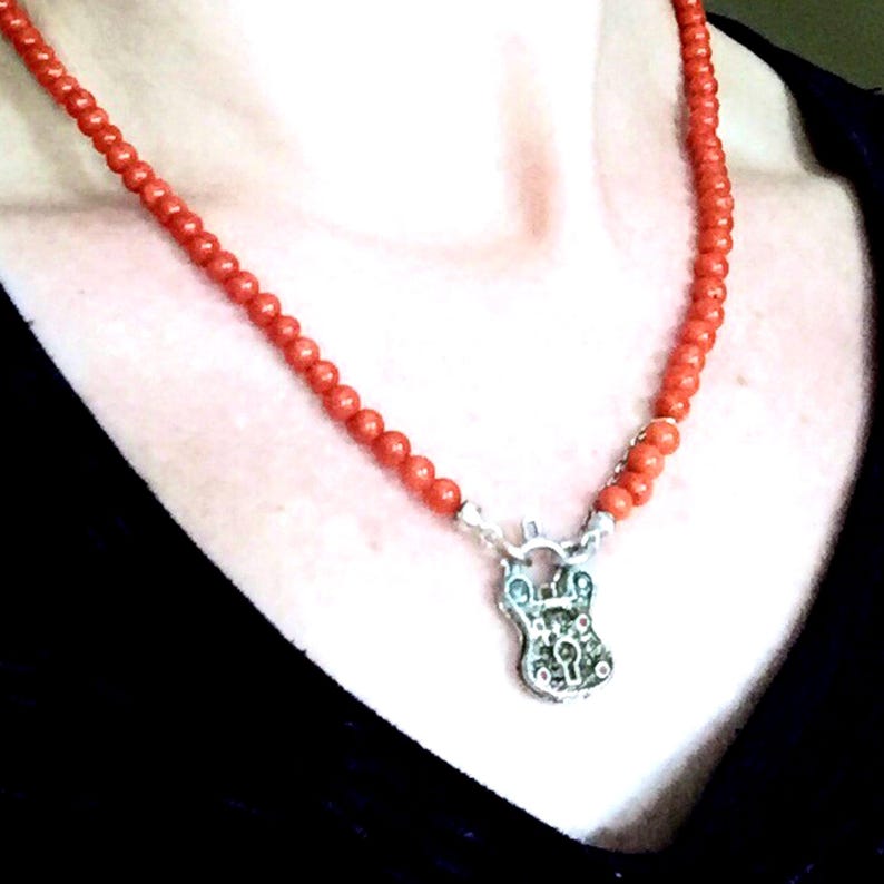 Puede incluir: Un collar de coral rojo con un colgante de candado de plata. El collar est&aacute; hecho de peque&ntilde;as cuentas redondas y el candado est&aacute; decorado con dise&ntilde;os intrincados.
