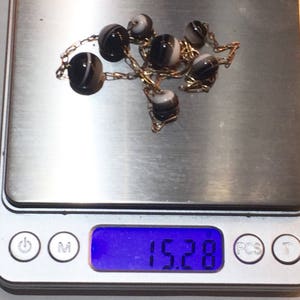 May include: A delicate necklace featuring alternating black and white striped beads strung on a gold chain. The necklace is displayed on a digital scale, with a weight reading of 15.28.