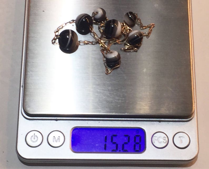 May include: A delicate necklace featuring alternating black and white striped beads strung on a gold chain. The necklace is displayed on a digital scale, with a weight reading of 15.28.