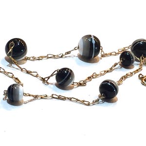 May include: A necklace featuring black and white striped beads strung on a delicate gold-colored chain. The beads are spherical and vary slightly in size. The chain has a classic link design, and the necklace is displayed on a white surface.