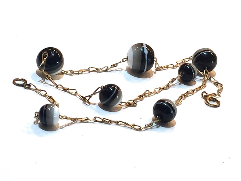 May include: A necklace featuring black and white striped beads strung on a delicate gold-colored chain. The beads are spherical and vary slightly in size. The chain has a classic link design, and the necklace is displayed on a white surface.