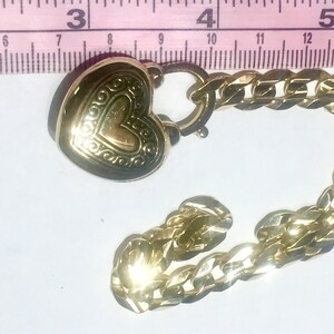 May include: A gold heart-shaped charm with an ornate design, attached to a gold chain. The chain is a curb style with a lobster clasp.