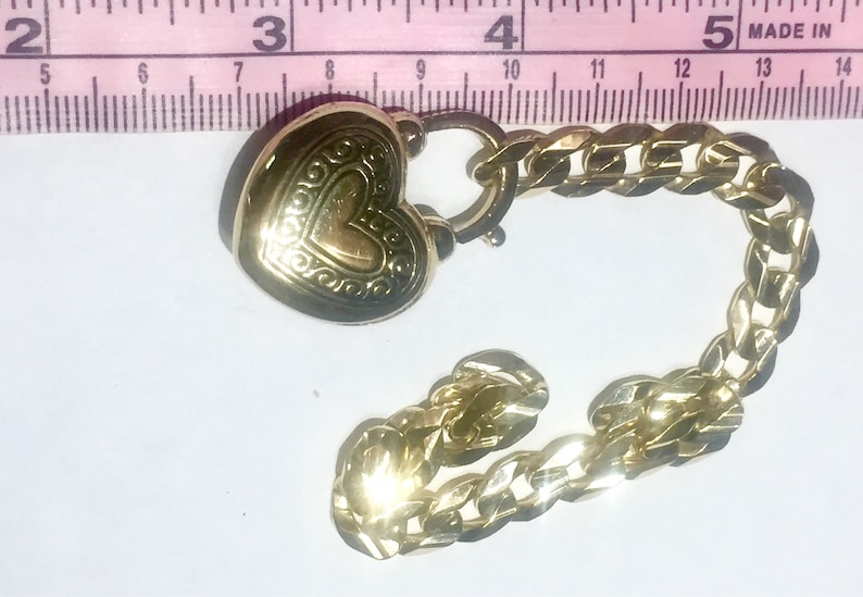 May include: A gold heart-shaped charm with an ornate design, attached to a gold chain. The chain is a curb style with a lobster clasp.