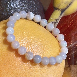 May include: A light blue beaded bracelet with round beads. The bracelet is resting on a yellow grapefruit. Other fruits are in the background.