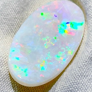 Puede incluir: Una piedra preciosa de ópalo pulida, de forma ovalada. El ópalo tiene una base blanca lechosa con destellos iridiscentes de verde, azul, rosa y amarillo. La gema se encuentra sobre un fondo de tela de color neutro.