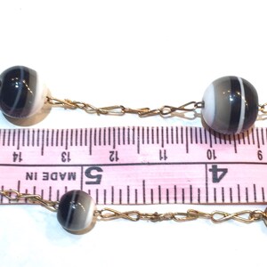 May include: A vintage necklace featuring striped, spherical beads in shades of black, white, and gray, strung on a delicate gold-tone chain. The beads are approximately 0.5 inches in diameter. The necklace is displayed against a white background with a pink ruler.