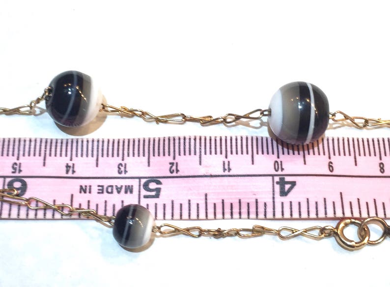 May include: A vintage necklace featuring striped, spherical beads in shades of black, white, and gray, strung on a delicate gold-tone chain. The beads are approximately 0.5 inches in diameter. The necklace is displayed against a white background with a pink ruler.
