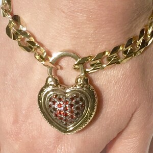 May include: A gold chain bracelet with a heart-shaped padlock charm. The padlock is decorated with red gemstones.