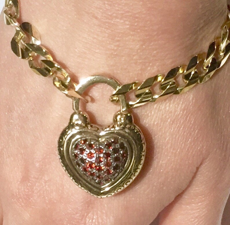 May include: A gold chain bracelet with a heart-shaped padlock charm. The padlock is decorated with red gemstones.