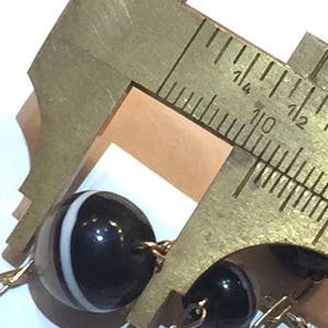 May include: Close-up of a black and white beaded necklace with a gold chain. A brass caliper is measuring the diameter of one of the beads. The beads are round and have a glossy finish. The caliper is marked with inch measurements.