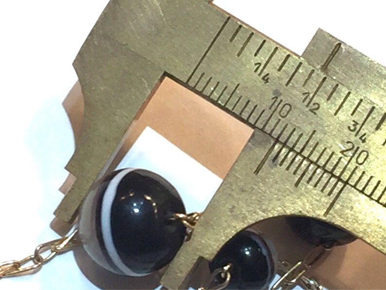 May include: Close-up of a black and white beaded necklace with a gold chain. A brass caliper is measuring the diameter of one of the beads. The beads are round and have a glossy finish. The caliper is marked with inch measurements.