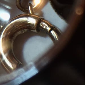 May include: Close-up of a gold-colored jewelry clasp. The clasp is curved and has a small, hinged section for opening and closing. The metal has a polished finish, reflecting light.