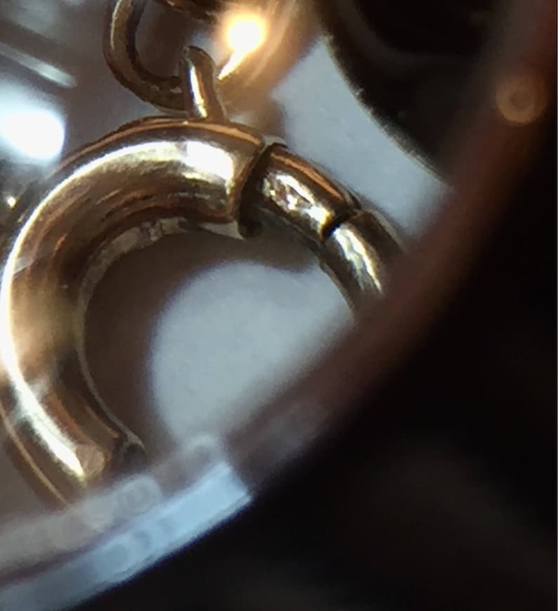 May include: Close-up of a gold-colored jewelry clasp. The clasp is curved and has a small, hinged section for opening and closing. The metal has a polished finish, reflecting light.