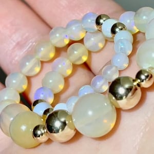 May include: Close-up of a bracelet featuring iridescent, round beads in shades of white and yellow. Gold-colored metal beads are interspersed throughout the bracelet. The bracelet is likely made of gemstones.