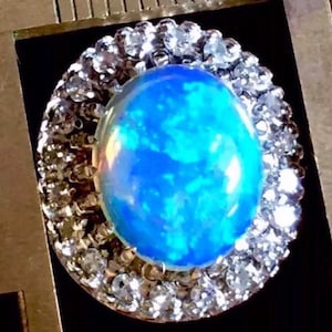 May include: A blue opal gemstone ring with a halo of clear round stones. The opal has a blue, green, and white iridescent sheen.