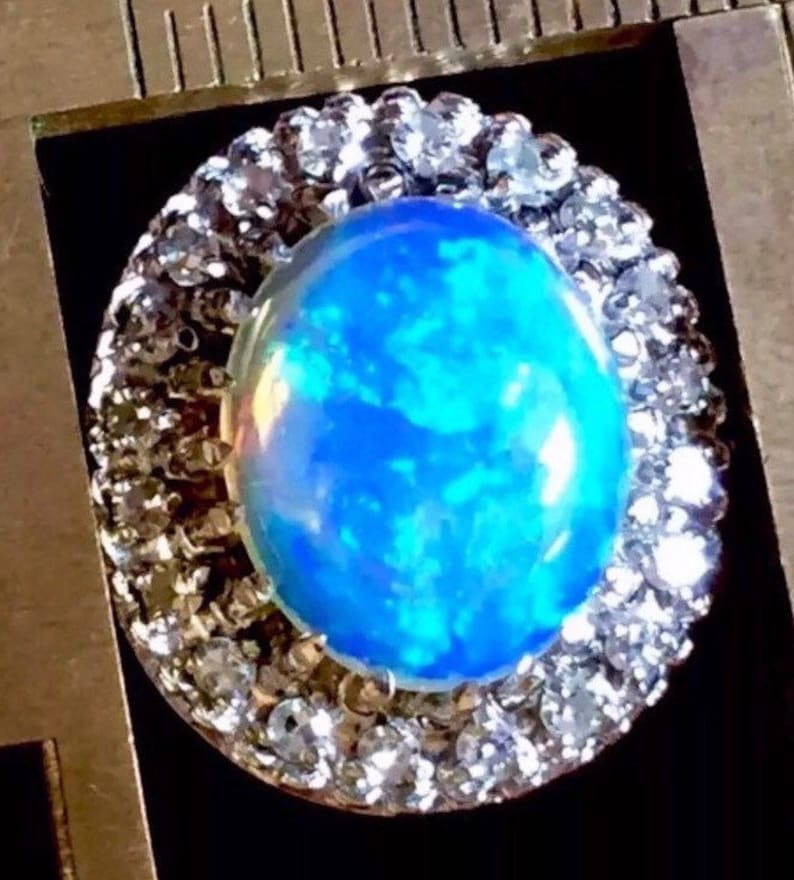 May include: A blue opal gemstone ring with a halo of clear round stones. The opal has a blue, green, and white iridescent sheen.