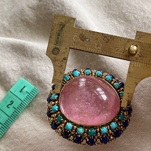 May include: A pink gemstone ring with a gold setting and a border of blue and turquoise stones. The ring is being measured with a ruler, showing it is approximately 2 cm in diameter.