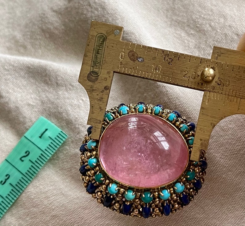 May include: A pink gemstone ring with a gold setting and a border of blue and turquoise stones. The ring is being measured with a ruler, showing it is approximately 2 cm in diameter.