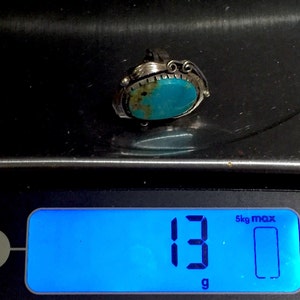 以下が含まれることがあります： 銀色の枠にセットされたターコイズの指輪。指輪は13グラムを示すデジタルスケールに乗っています。