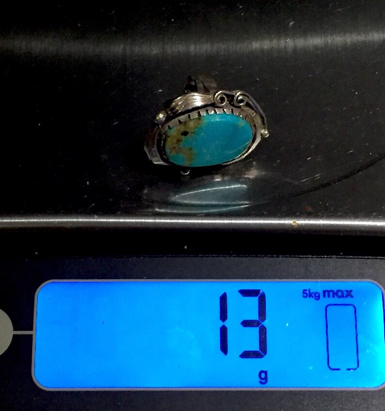 以下が含まれることがあります： 銀色の枠にセットされたターコイズの指輪。指輪は13グラムを示すデジタルスケールに乗っています。