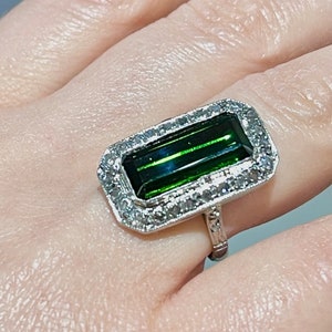 May include: A silver ring featuring a rectangular, emerald-green gemstone with a halo of small, clear stones. The ring's band has a detailed, ornate design. The gemstone's color is a deep, rich green.