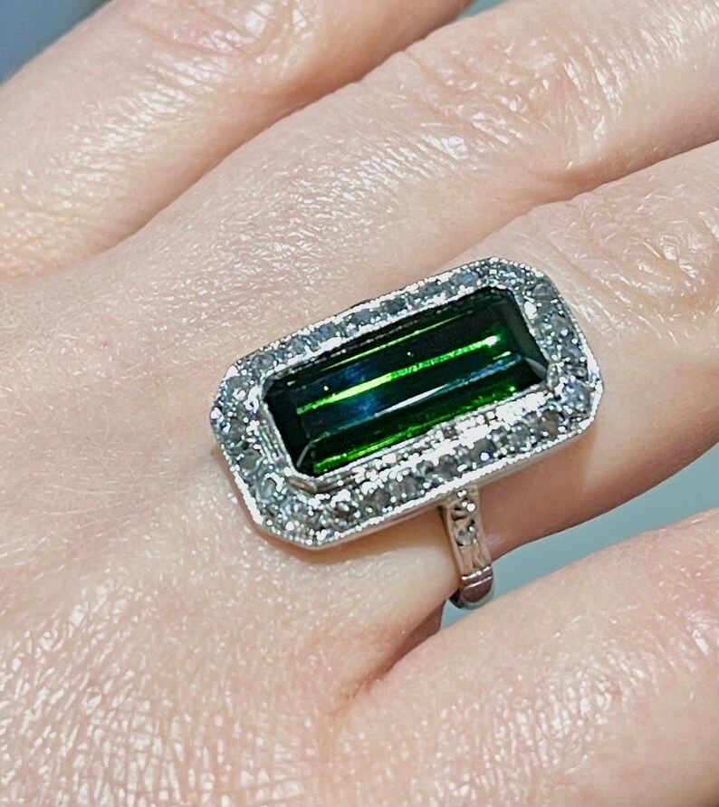 May include: A silver ring featuring a rectangular, emerald-green gemstone with a halo of small, clear stones. The ring's band has a detailed, ornate design. The gemstone's color is a deep, rich green.