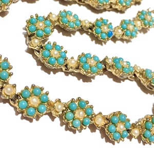 Puede incluir: Un collar de metal dorado con un patrón floral repetitivo. Cada flor consta de seis pequeñas cuentas redondas de color turquesa brillante que rodean una sola cuenta central de color crema. Los eslabones están unidos en una cadena continua.