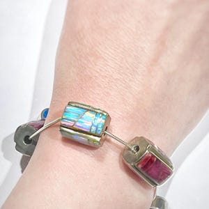 May include: A silver bracelet with two colorful, rectangular beads. The beads are made of inlaid stone and feature a variety of colors, including blue, pink, and red.
