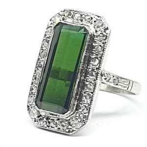 May include: A rectangular, emerald-cut green gemstone ring with a silver band. The gemstone is surrounded by a halo of small, clear stones. The ring has a vintage or antique style.