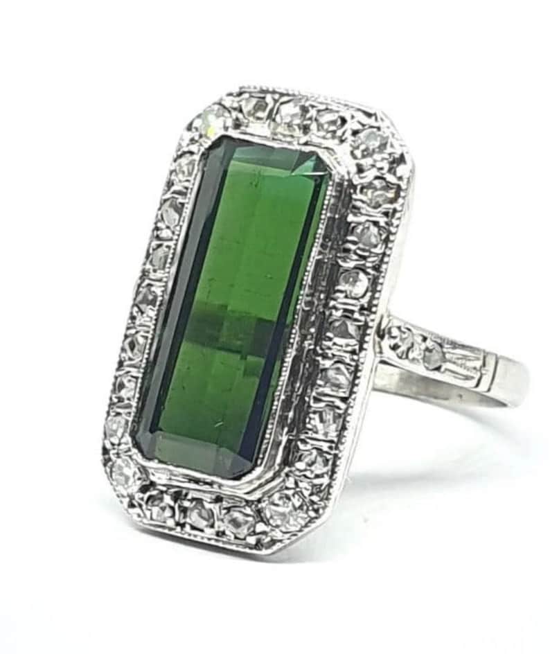May include: A rectangular, emerald-cut green gemstone ring with a silver band. The gemstone is surrounded by a halo of small, clear stones. The ring has a vintage or antique style.