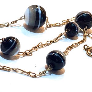 May include: A gold-toned chain necklace featuring alternating black and white striped beads and solid black beads. The beads are round and connected by a delicate chain, creating an elegant and classic jewelry piece.