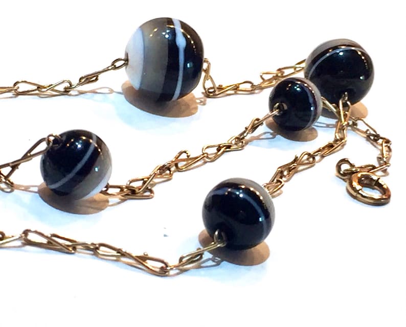 May include: A gold-toned chain necklace featuring alternating black and white striped beads and solid black beads. The beads are round and connected by a delicate chain, creating an elegant and classic jewelry piece.