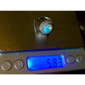 May include: A silver ring with a large opal gemstone surrounded by smaller white gemstones. The ring is on a digital scale that reads 5.83.
