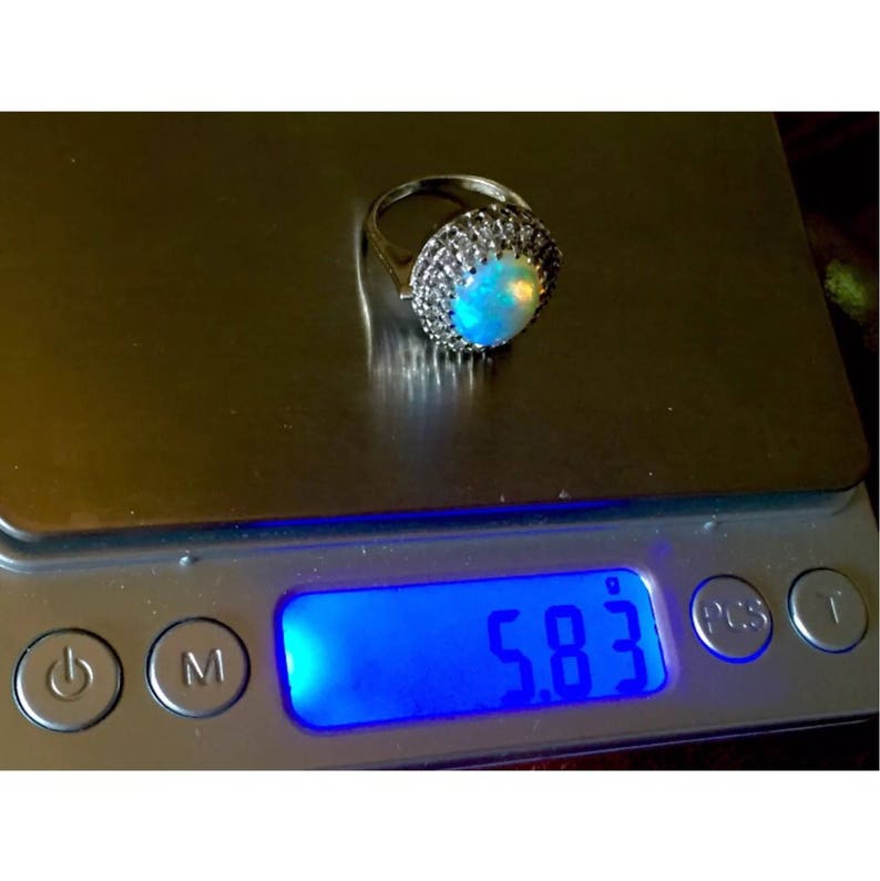 May include: A silver ring with a large opal gemstone surrounded by smaller white gemstones. The ring is on a digital scale that reads 5.83.