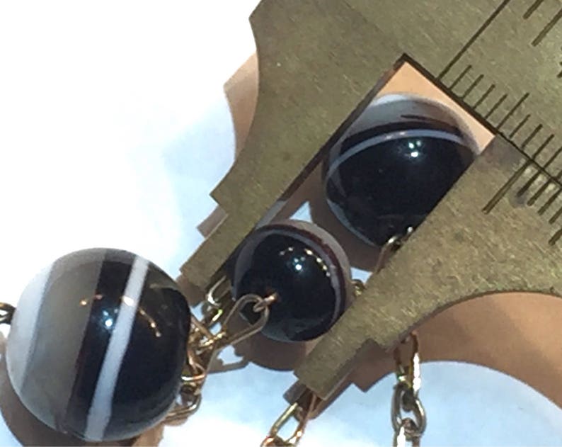 May include: Close-up of a gold chain with several round beads. The beads are black, white, and gray with striped patterns. A brass ruler is used to measure the beads. The background is white.