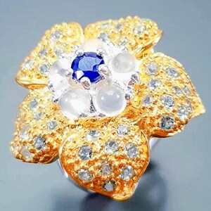 May include: A flower-shaped ring featuring a central deep blue round gemstone surrounded by four smooth, translucent white cabochon stones. The five petals are textured with a gold finish and are encrusted with numerous small, clear white stones.